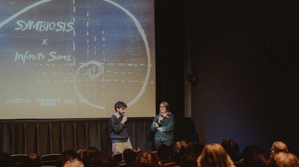 A photo of Sonya Hansen and Diego Murillo at the Symbiosis film screening, standing in front of a screen that says Symbiosis x Infinite Sums.