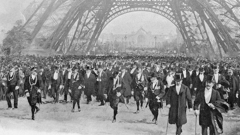 Many gentlemen in top hats and black suits underneath the Eiffel Tower.
